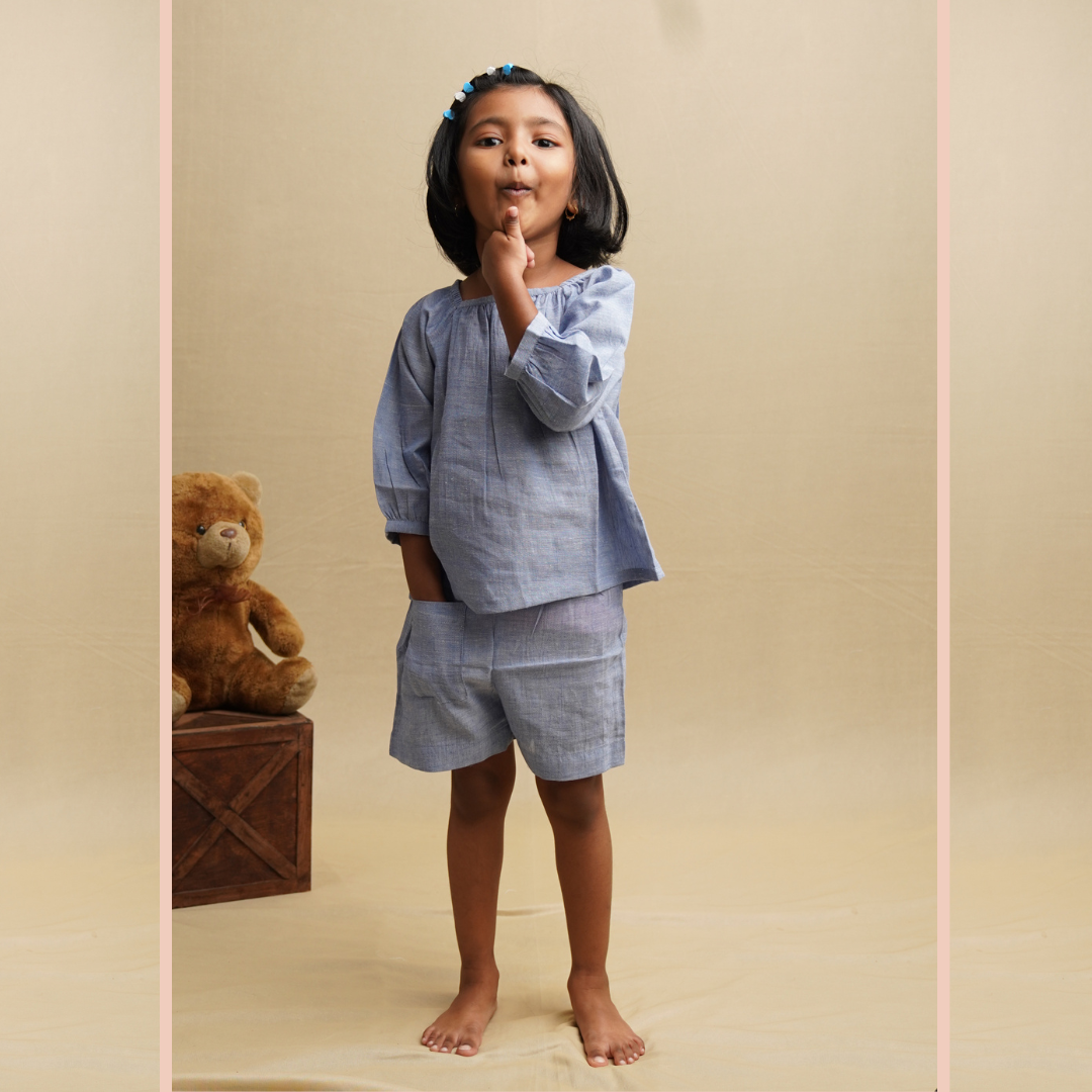 A girl wearing a blue chambray co-ord set with puff sleeves and shorts, standing against a beige background