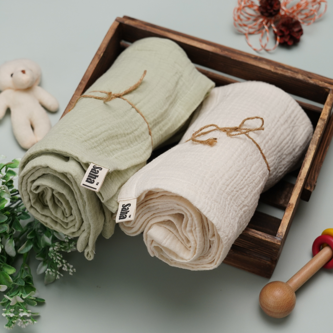 Two rolled-up muslin swaddle wraps, one green and one beige, in a wooden crate