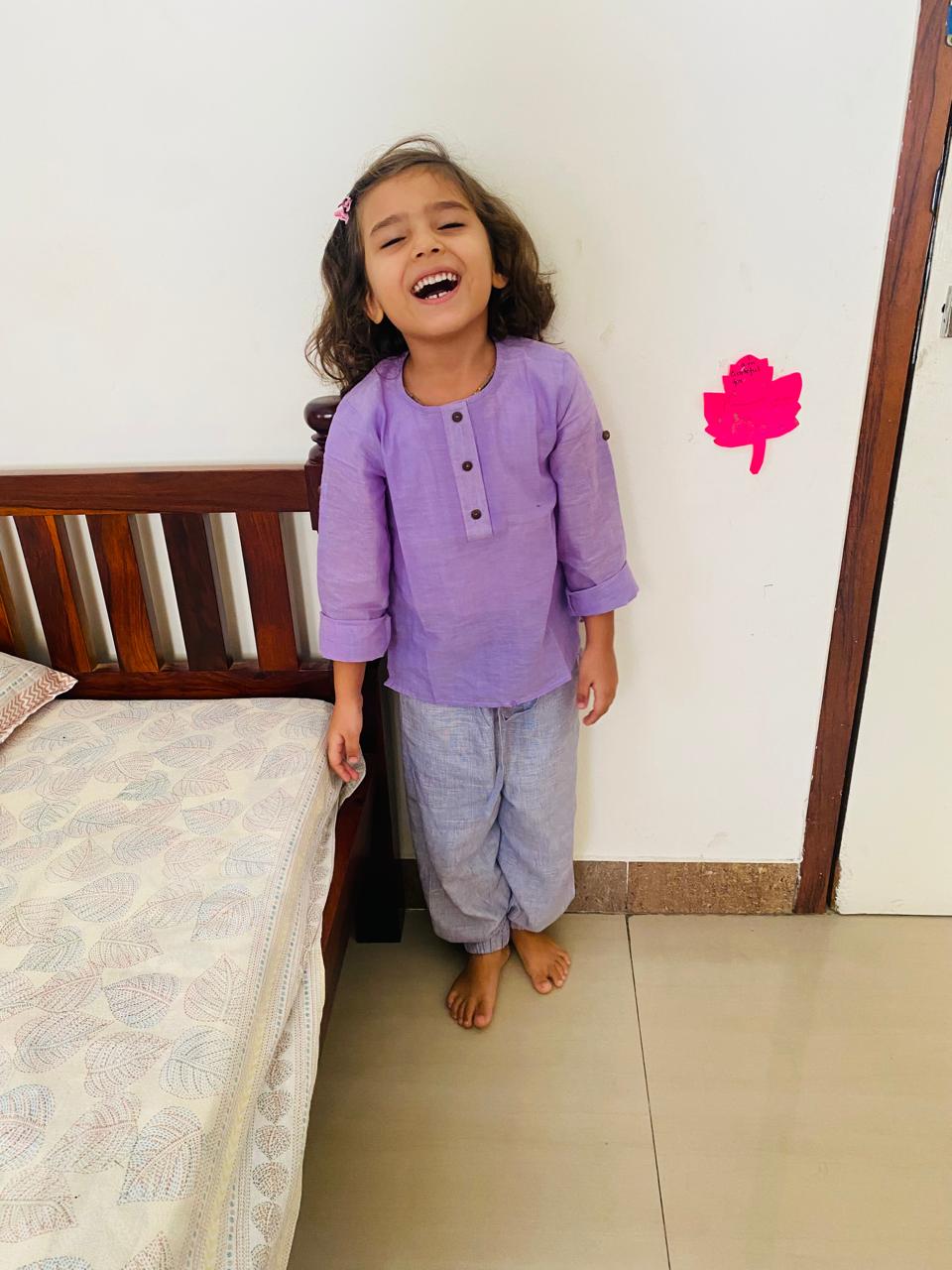 Child in a pure cotton purple shirt and pure cotton purple jogger pants standing in a room with a bed and wall decorations.