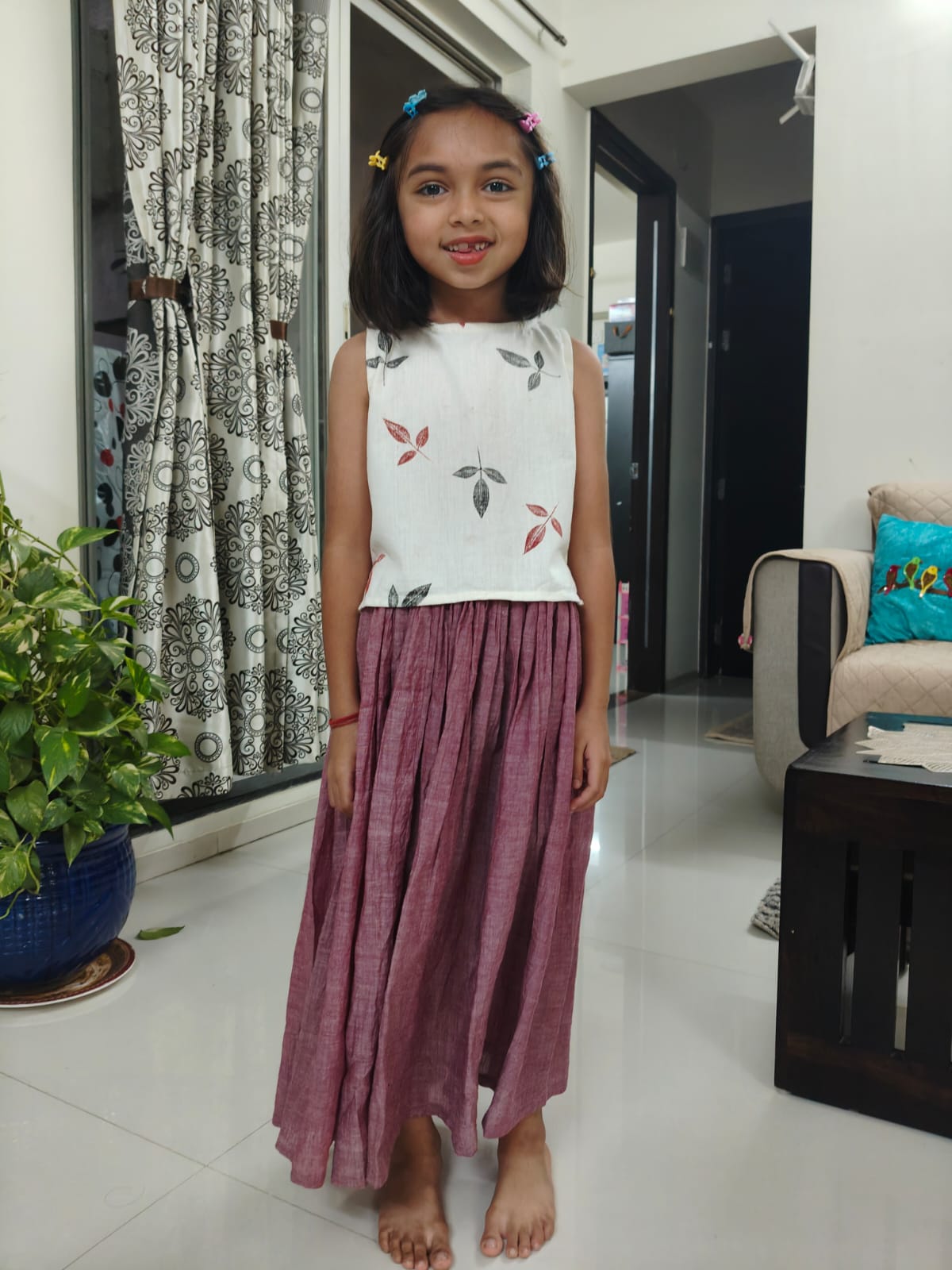 Young girl standing in a living room wearing a white top with floral patterns and a long cotton skirt.