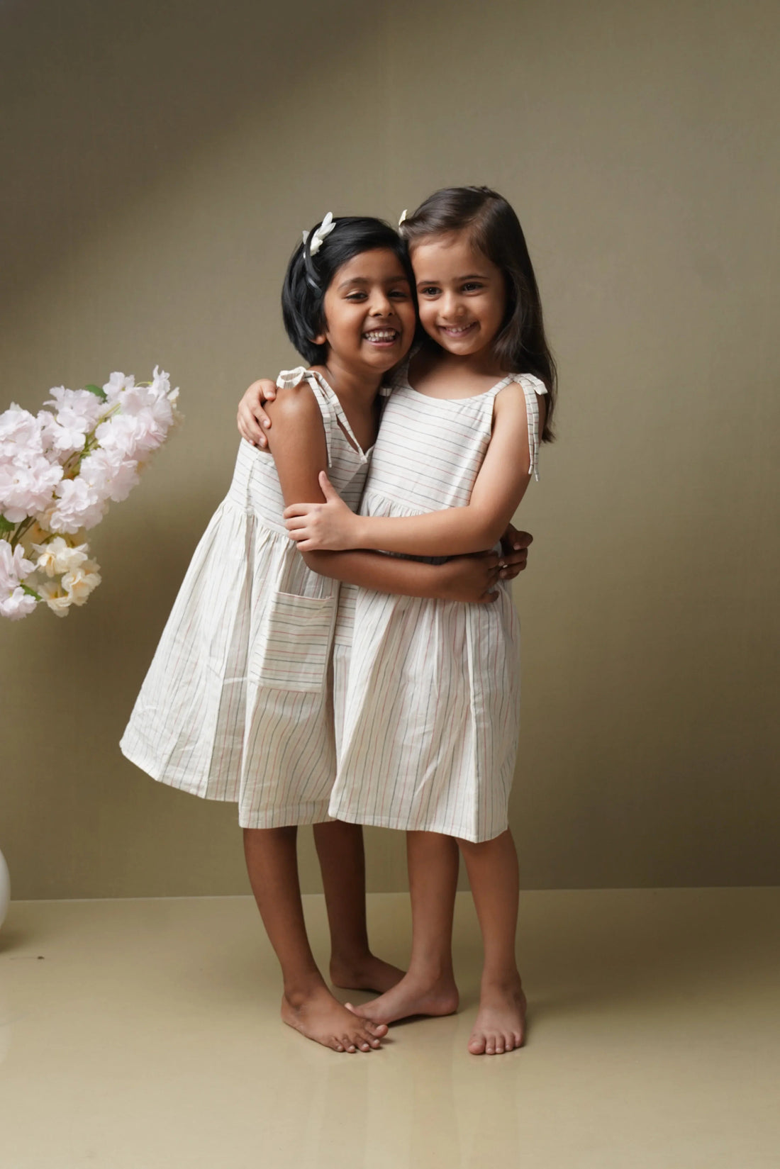 Two smiling girls in matching striped organic cotton dresses, hugging in a minimalist room