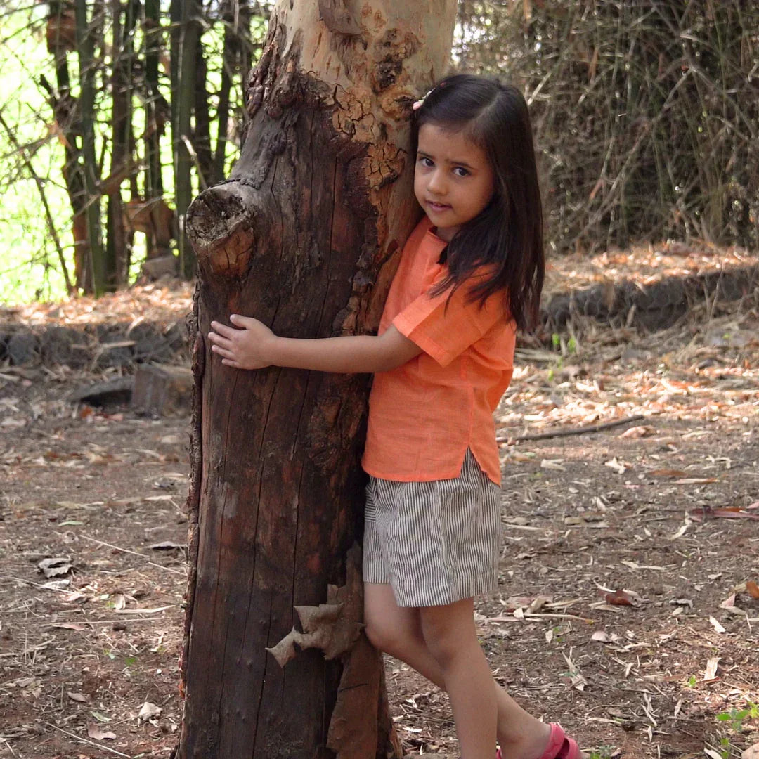 Girl wearing orange cotton top and striped shorts hugging a tree outdoors in summer