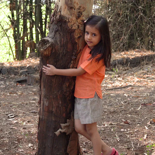 Girl wearing orange cotton top and striped shorts hugging a tree outdoors in summer