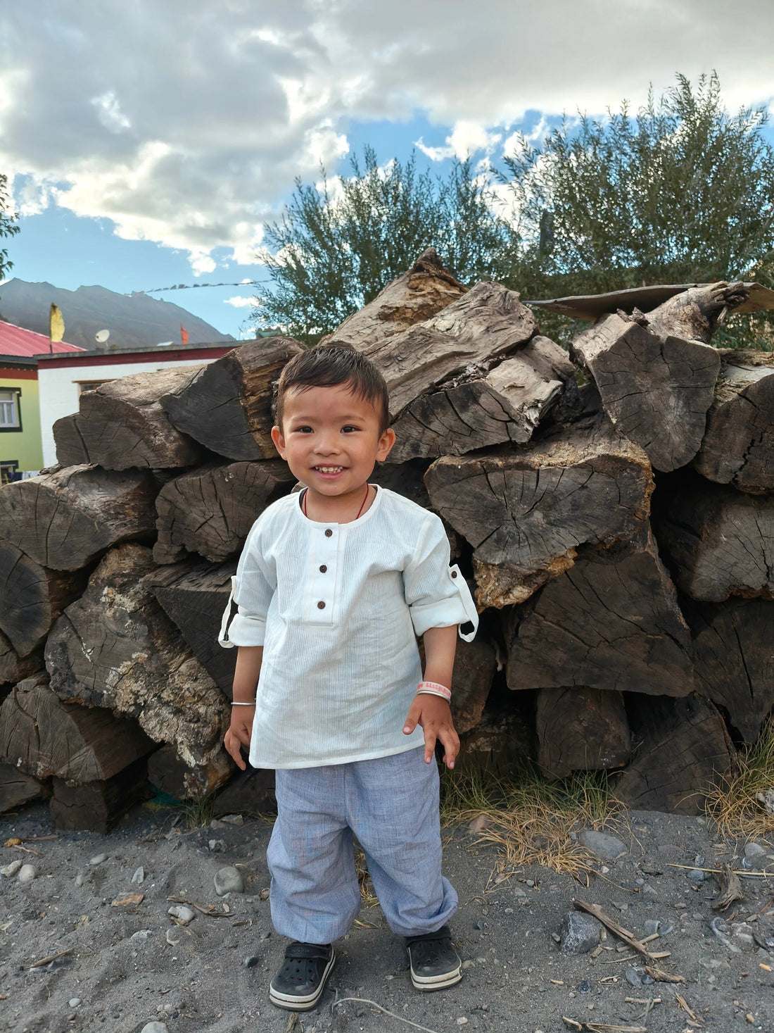Smiling child in organic khadi cotton baby clothes standing outdoors in front of stacked logs