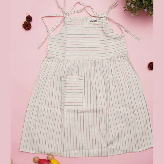 White cotton striped dress for girls with front pocket, tie straps, laid on pink background