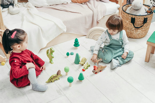 Two young children playing on the floor with wooden dinosaur toys and trees in a cozy kids' room.