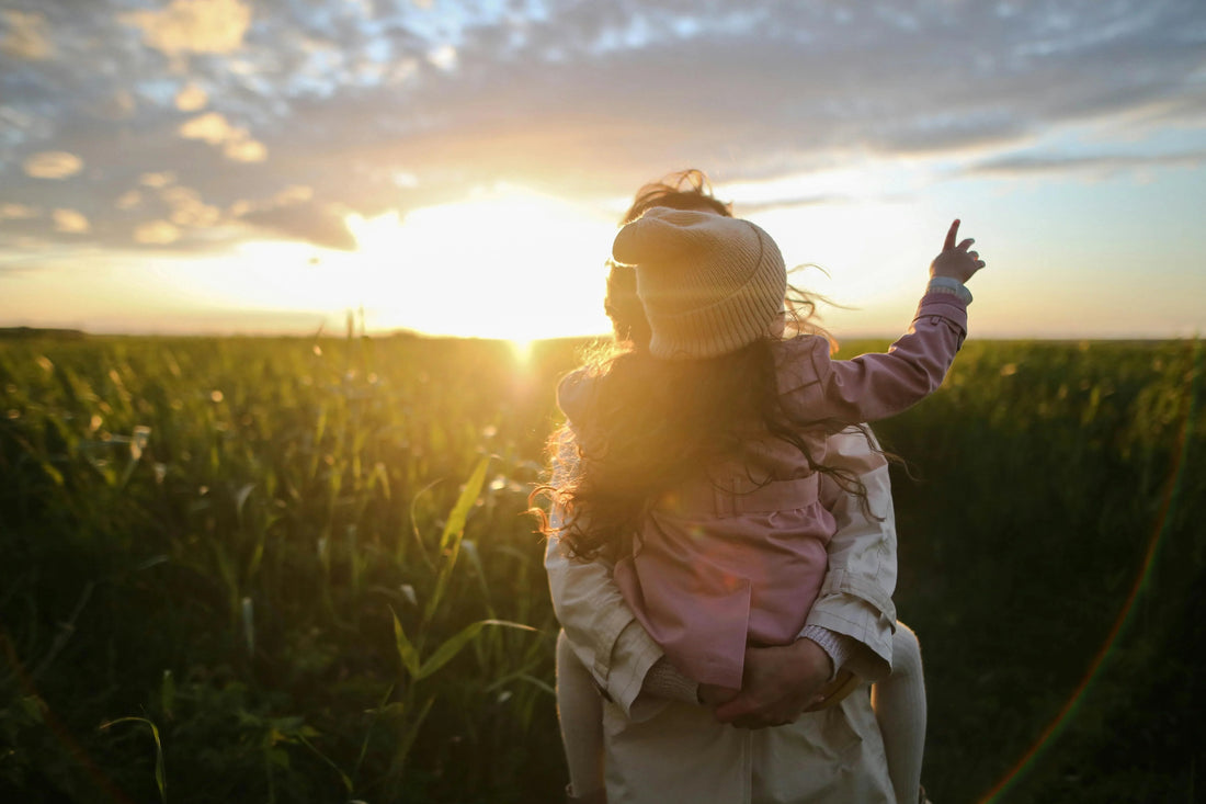 Mother holding child in organic cotton clothing in a green field at sunset