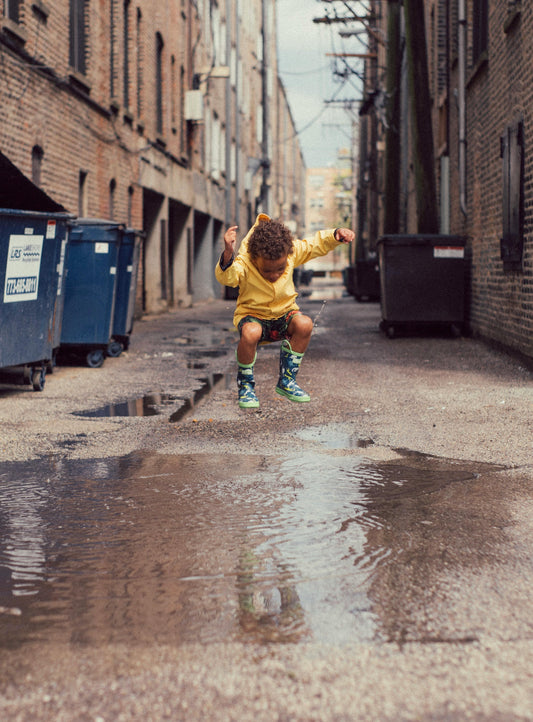 Child in khadi raincoat and boots jumping in puddle on urban street, playful kids fashion