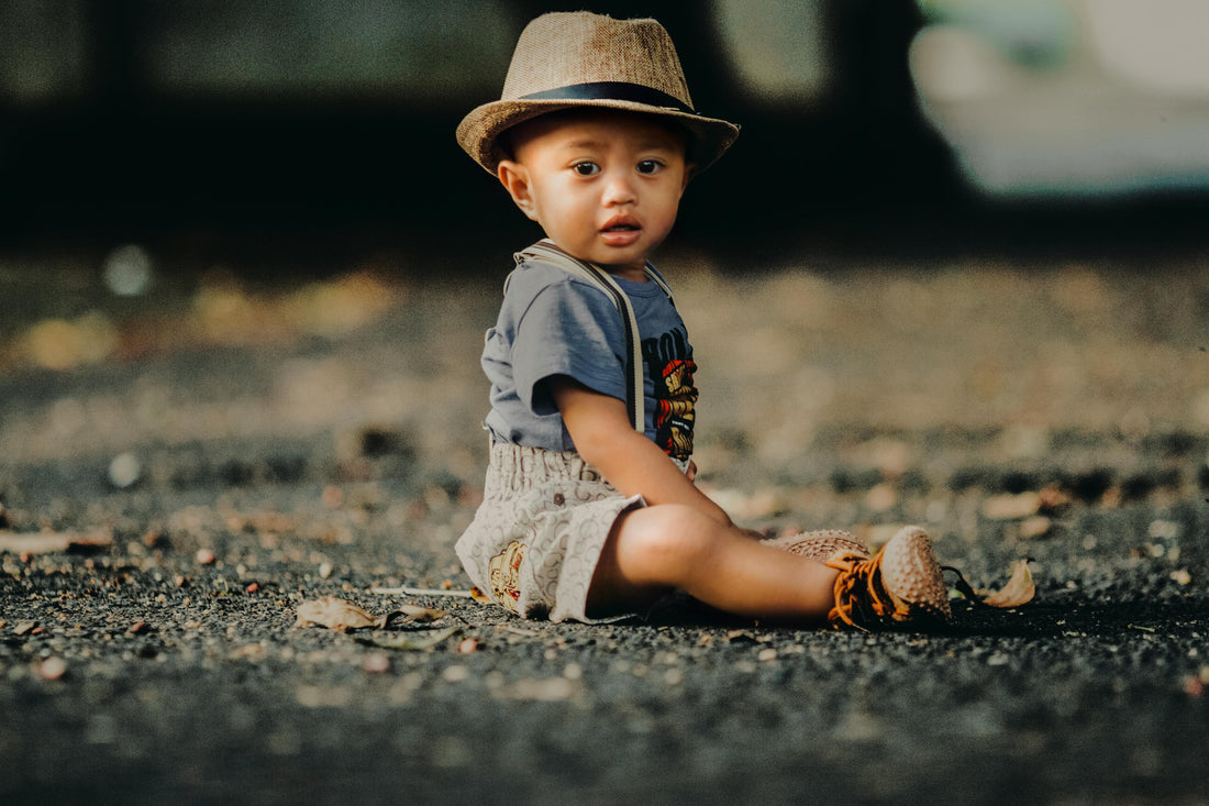 Baby boy in khadi shorts, grey t-shirt, and straw hat sitting outdoors