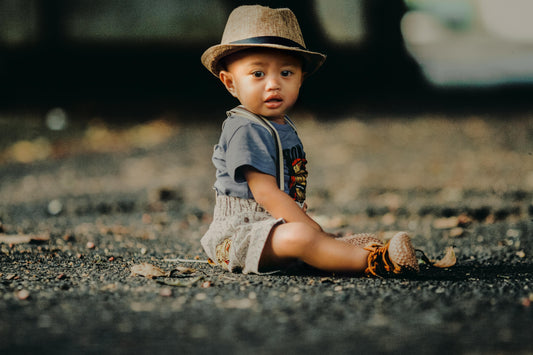 Baby boy in khadi shorts, grey t-shirt, and straw hat sitting outdoors