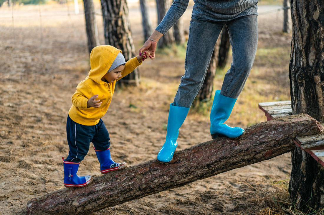 Toddler in yellow hoodie and blue rain boots balancing on log with adult outdoors
