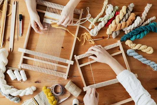 Hands weaving with wooden looms, colorful yarn, and natural fibers on a wooden table