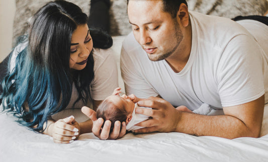 Parents in white shirts lying on bed gently holding newborn baby, family bonding moment