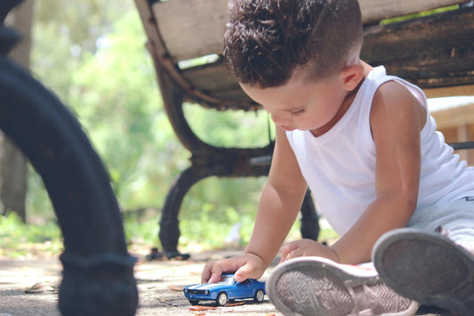Toddler in white tank top playing with blue toy car outdoors near a bench, wooden toys inspiration