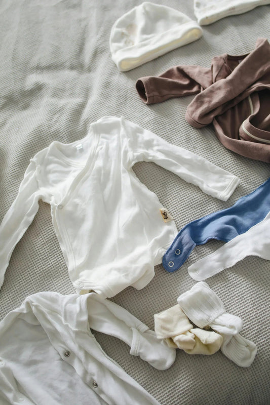 Organic cotton baby clothes and accessories, including white bodysuits, hats, and socks on a bed