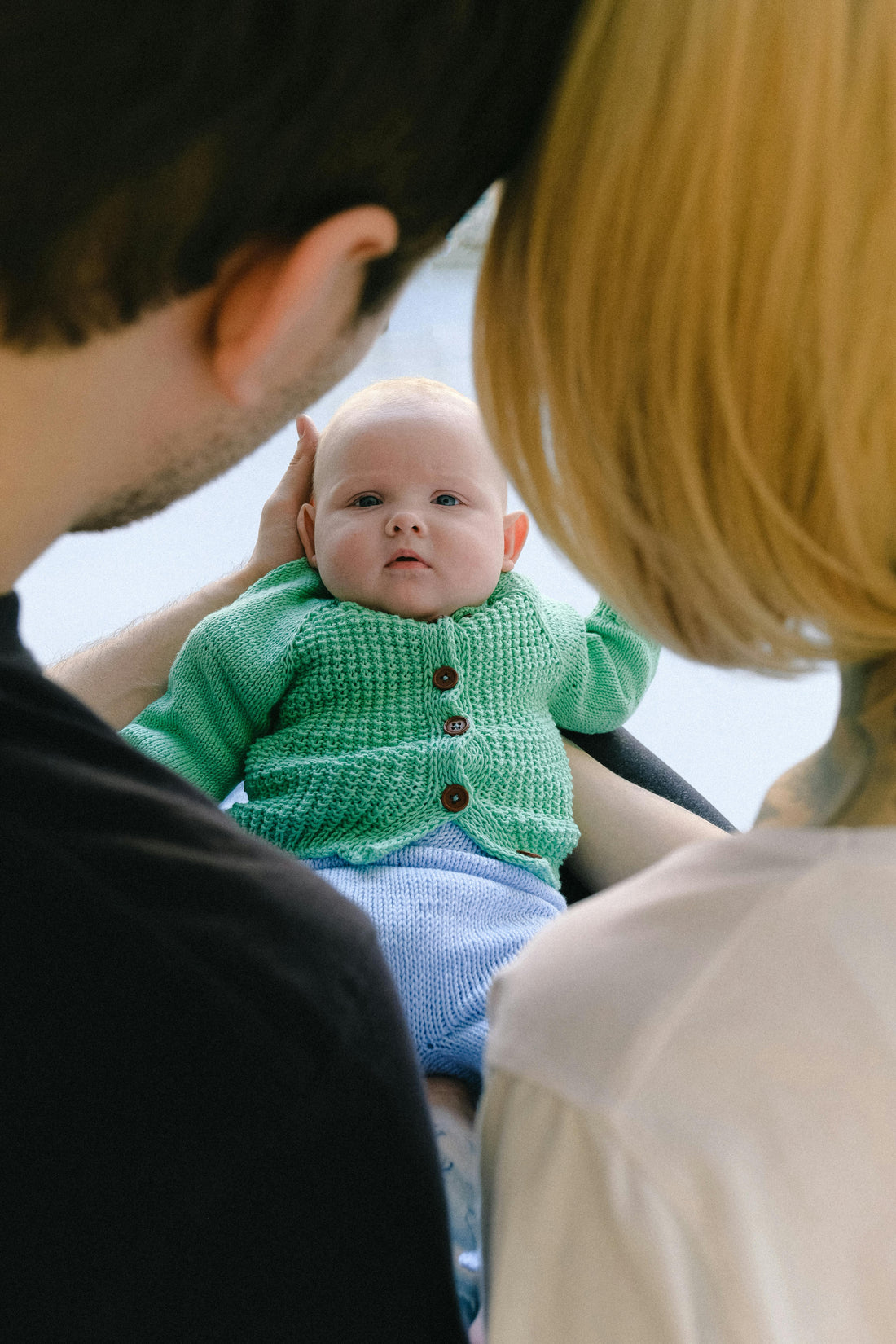 Baby in green knitted sweater and blue organic cotton pants held by adults