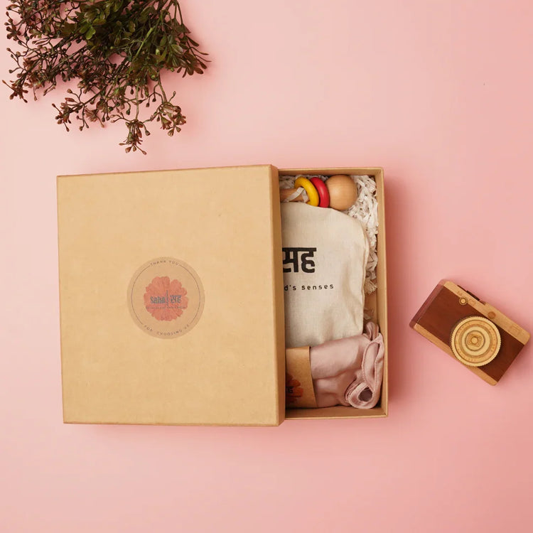 Gift box with wooden educational toys, fabric pouch, and baby clothes on pink background