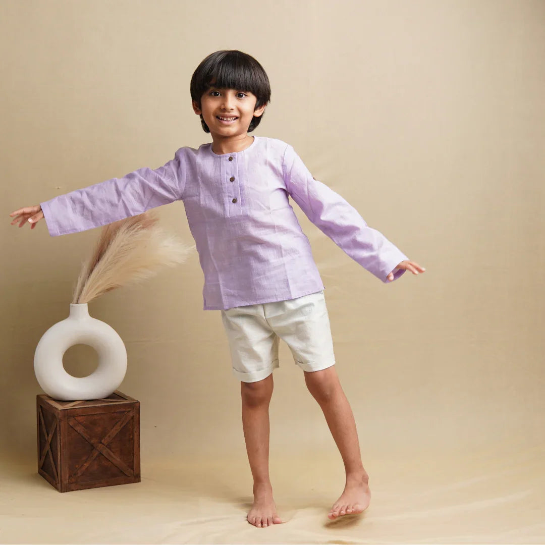Smiling boy in lavender khadi kurta and white shorts, barefoot, posing against beige background