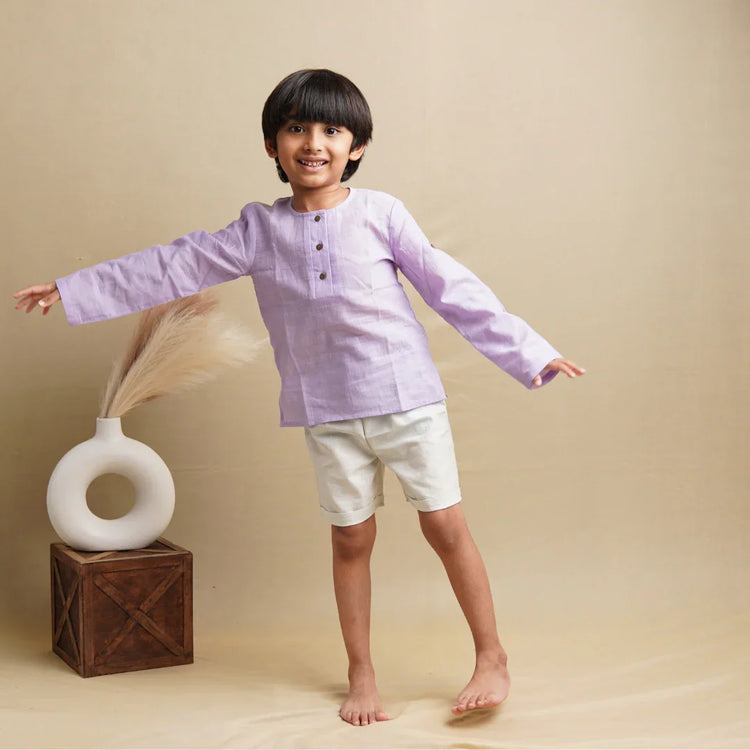 Smiling boy in lavender khadi kurta and white shorts, barefoot, posing against beige background