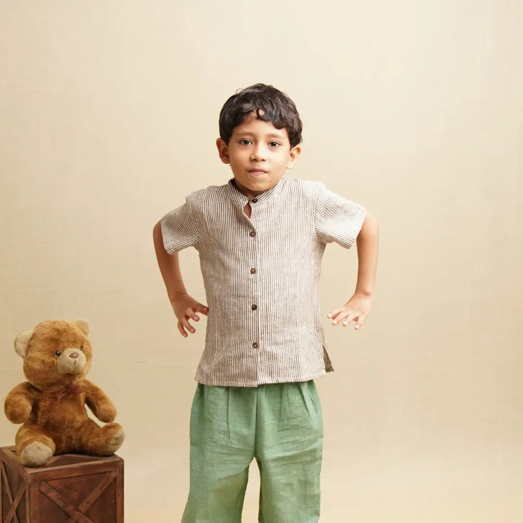 Child wearing a striped shirt and green pants standing next to a teddy bear on a beige background