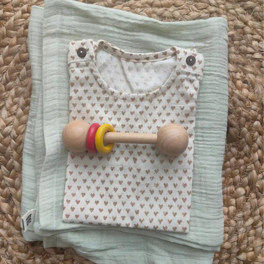 Folded baby outfit with heart pattern and wooden rattle on a muslin swaddle
