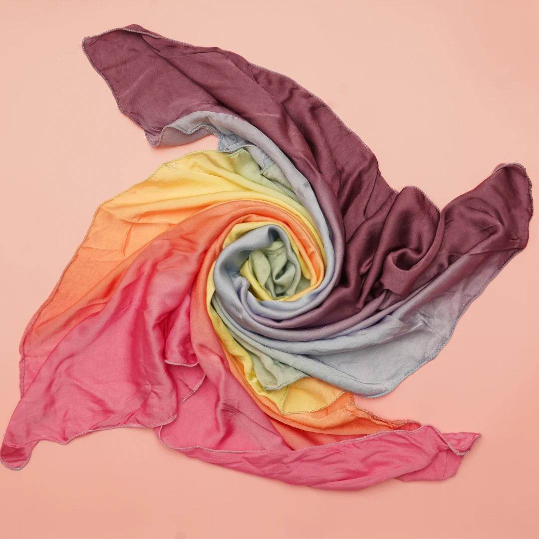 Hand-dyed soft rainbow semi-giant playsilk in pastel rainbow shades displayed in a spiral arrangement on a peach background