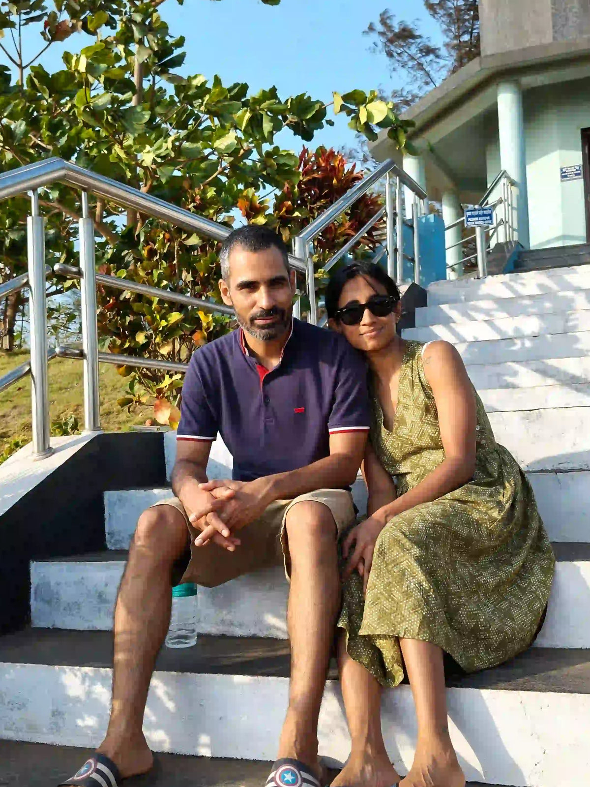 A man and a woman sitting together on outdoor concrete stairs in front of a building, with trees and a blue sky in the background. The man wears a dark blue polo shirt and beige shorts, and the woman wears a green sleeveless dress and sunglasses.