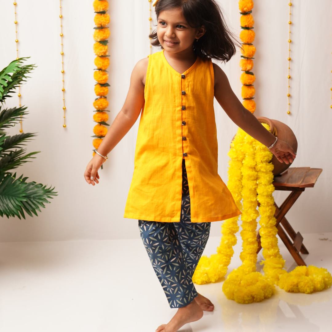 Young girl in a yellow top and patterned pants standing next to decorative yellow flowers.