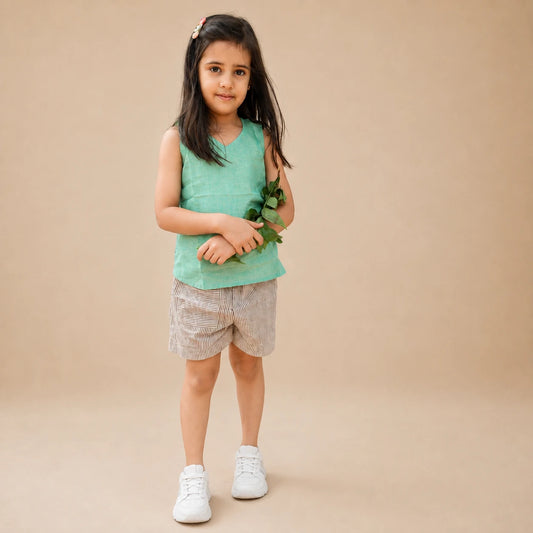 A young girl with long dark hair stands against a solid beige studio background, wearing a sleeveless mint green top, light beige patterned shorts, and white sneakers while holding a small green leafy sprig.