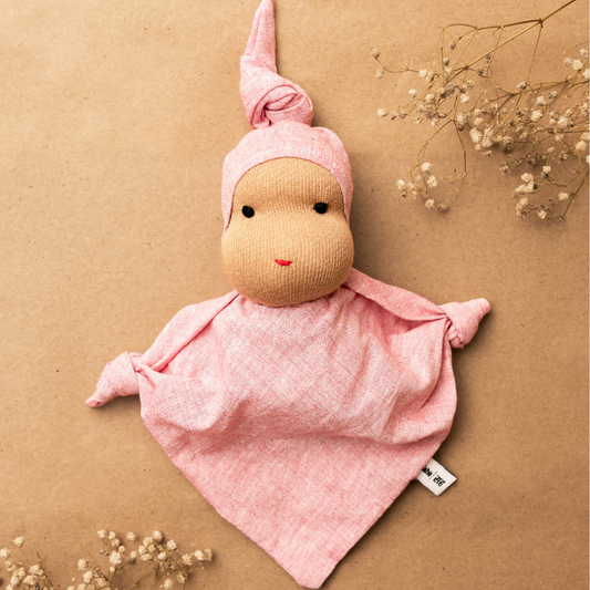 A handmade Waldorf fabric doll in pink, with a simple design and a knot at the top, displayed on a beige background with some floral elements.