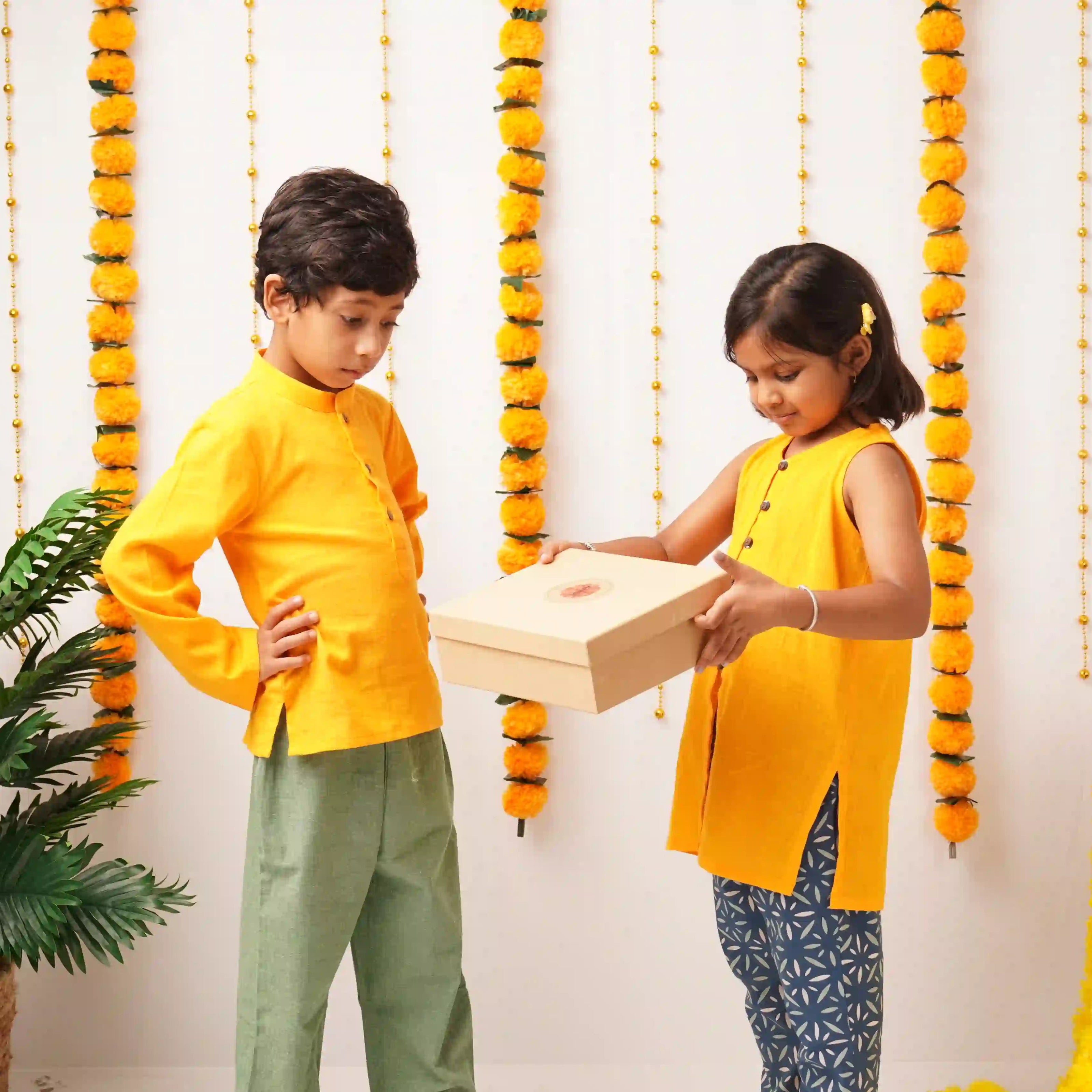 Two children wearing traditional Indian attire, one in a yellow kurta and green trousers and the other in a yellow sleeveless top and patterned pants, holding a gift box in front of a decorated wall with yellow marigold garlands.