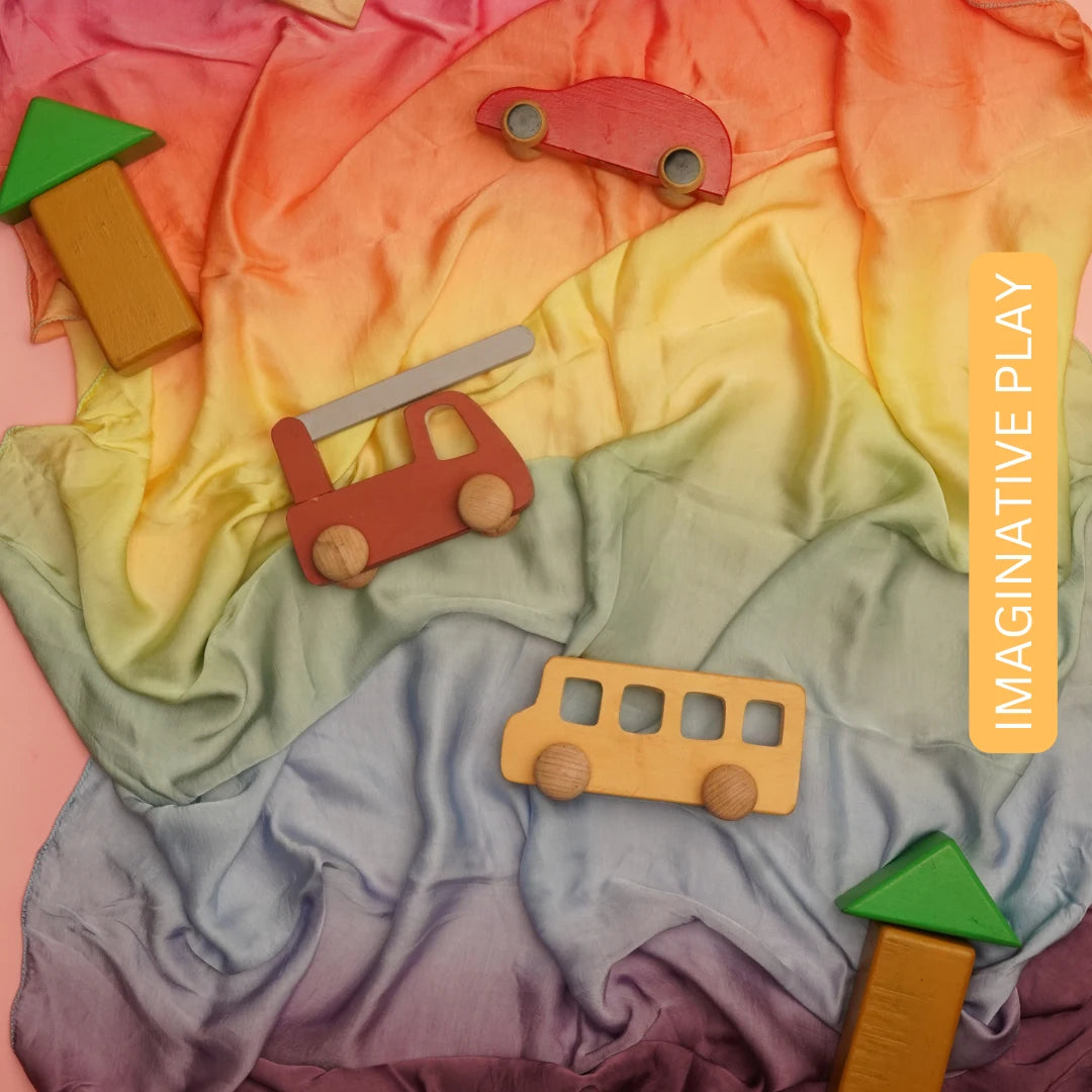 Wooden toy vehicles and blocks arranged on a soft rainbow semi-giant playsilk for open-ended imaginative play