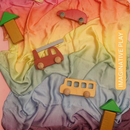 Wooden toy vehicles and blocks arranged on a soft rainbow semi-giant playsilk for open-ended imaginative play