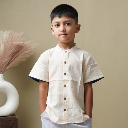 A young boy wearing a light beige linen short-sleeve shirt with dark navy blue inner collar and sleeve trim, standing against a neutral beige background.