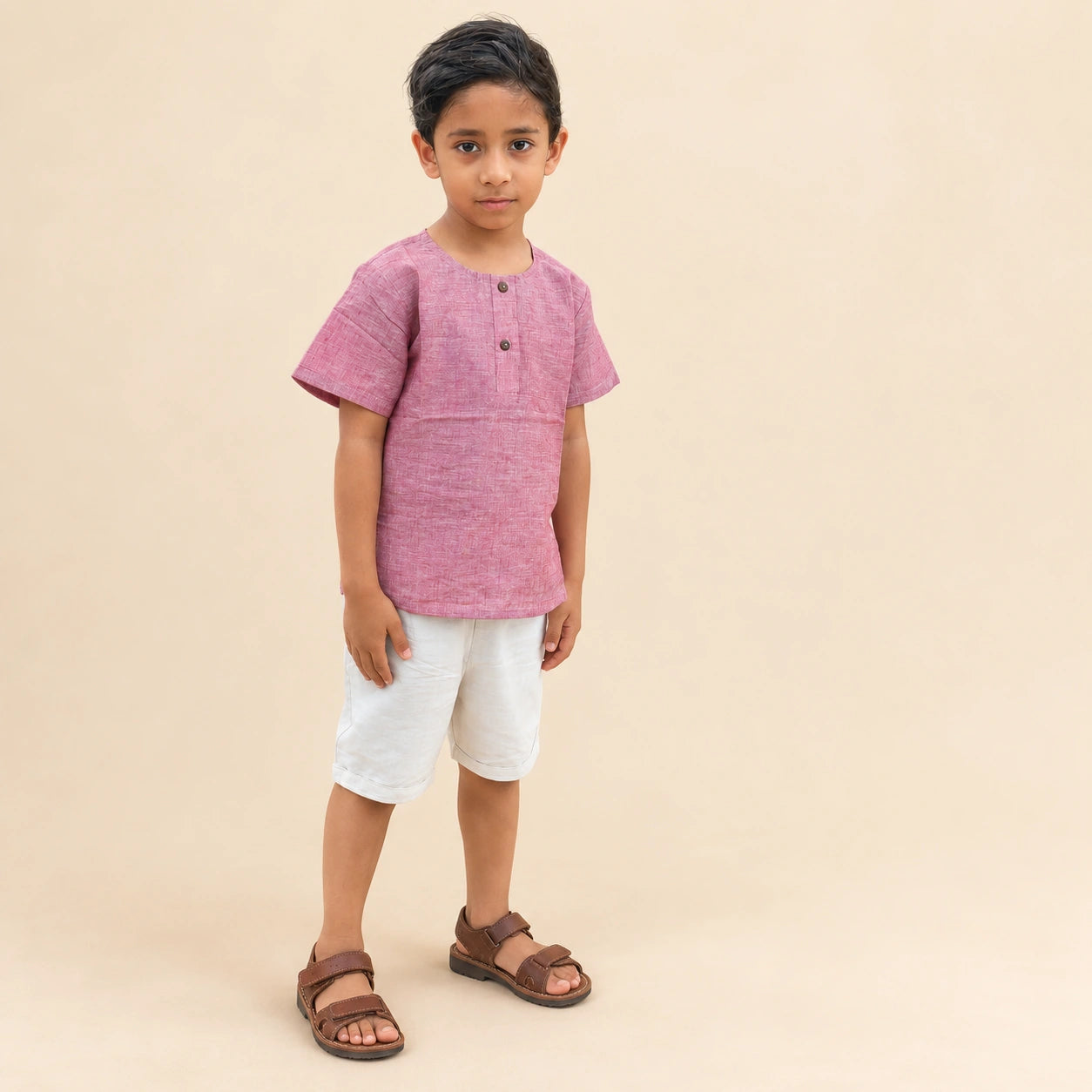 A young boy modeling a short-sleeve pink linen henley t-shirt, white linen shorts, and brown leather sandals against a solid beige studio background.