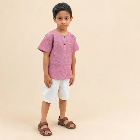 A young boy modeling a short-sleeve pink linen henley t-shirt, white linen shorts, and brown leather sandals against a solid beige studio background.