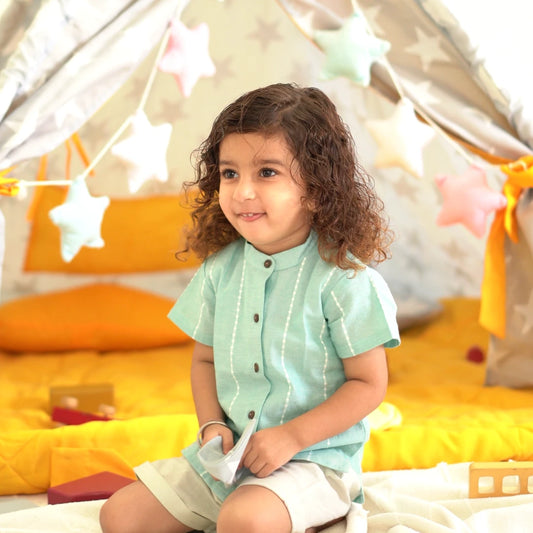 Child wearing a mint green handloom cotton shirt with beige shorts, sitting inside a cozy play tent with soft toys and cushions