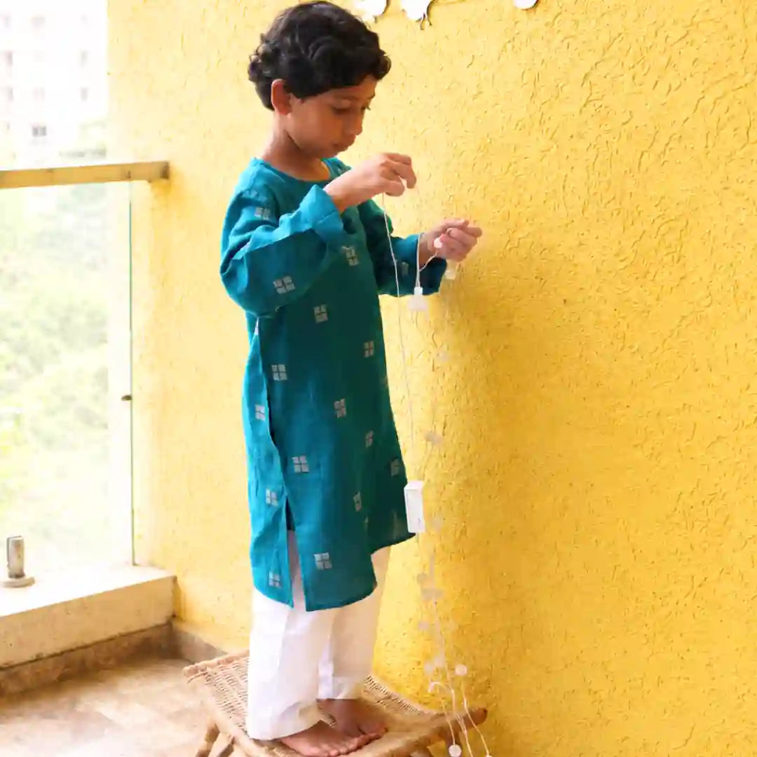 Boy in a teal handloom cotton kurta and white pajama standing on a stool, hanging festive lights on a yellow wall