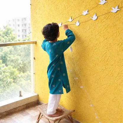 Boy in a teal handloom cotton kurta and white pajama hanging a paper bird garland on a yellow wall