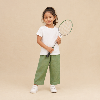 A young girl stands against a warm beige studio background holding a tennis racket. She is wearing a white short-sleeve t-shirt, sage green wide-leg pants, and white sneakers.