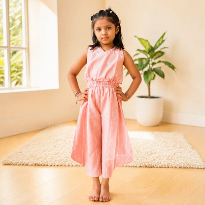 A young girl stands barefoot indoors wearing a sleeveless light pink jumpsuit with a gathered elastic waist and wide legs