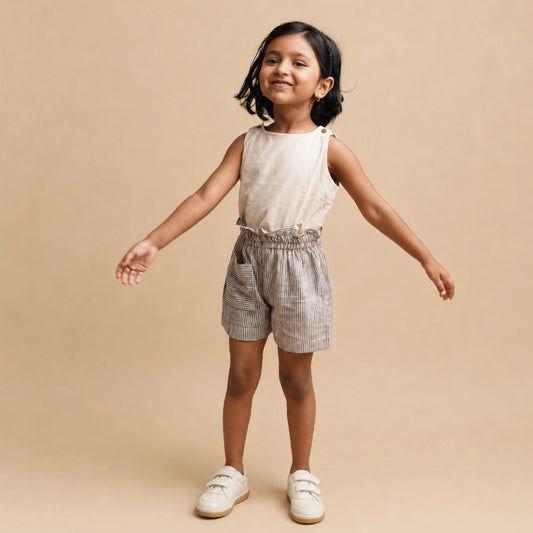 A young girl modeling a sleeveless beige top and gray-and-white striped shorts with a ruffled waistband, standing against a solid beige background.