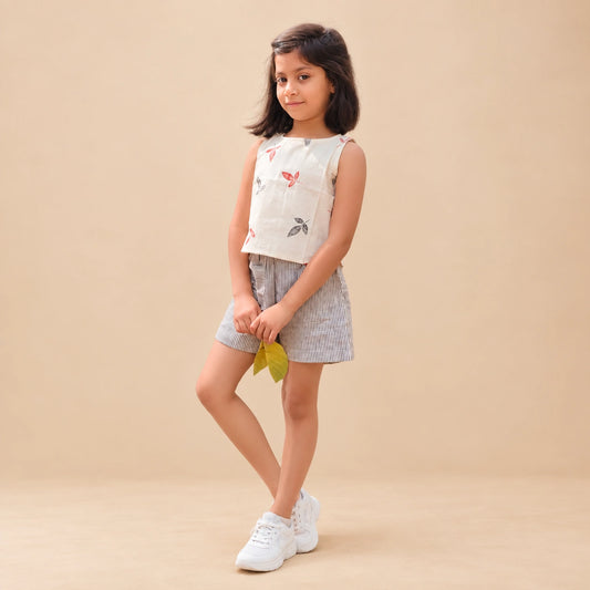 A young girl stands  wearing a sleeveless white top with small red and grey leaves prints