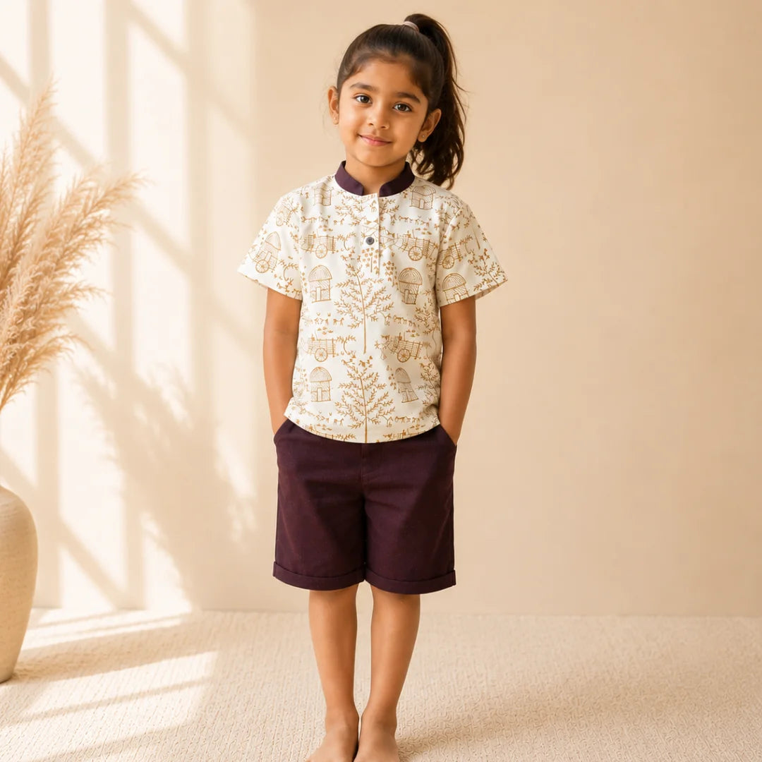 A young girl stands barefoot in a studio setting, wearing a short-sleeve button-up shirt with a cream base and tan botanical pattern, paired with dark plum shorts