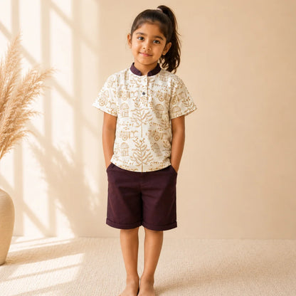 A young girl stands barefoot in a studio setting, wearing a short-sleeve button-up shirt with a cream base and tan botanical pattern, paired with dark plum shorts