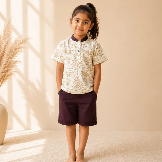 A young girl stands barefoot in a studio setting, wearing a short-sleeve button-up shirt with a cream base and tan botanical pattern, paired with dark plum shorts