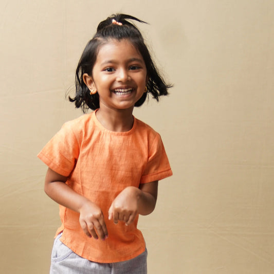 A young girl with her hair in a ponytail stands wearing a cream-colored short-sleeve kurta with a brown cart and tree pattern, paired with dark purple trousers. 
