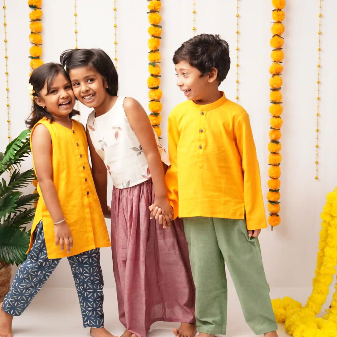Three children in colorful cotton festivewear standing together with decorative elements in the background.