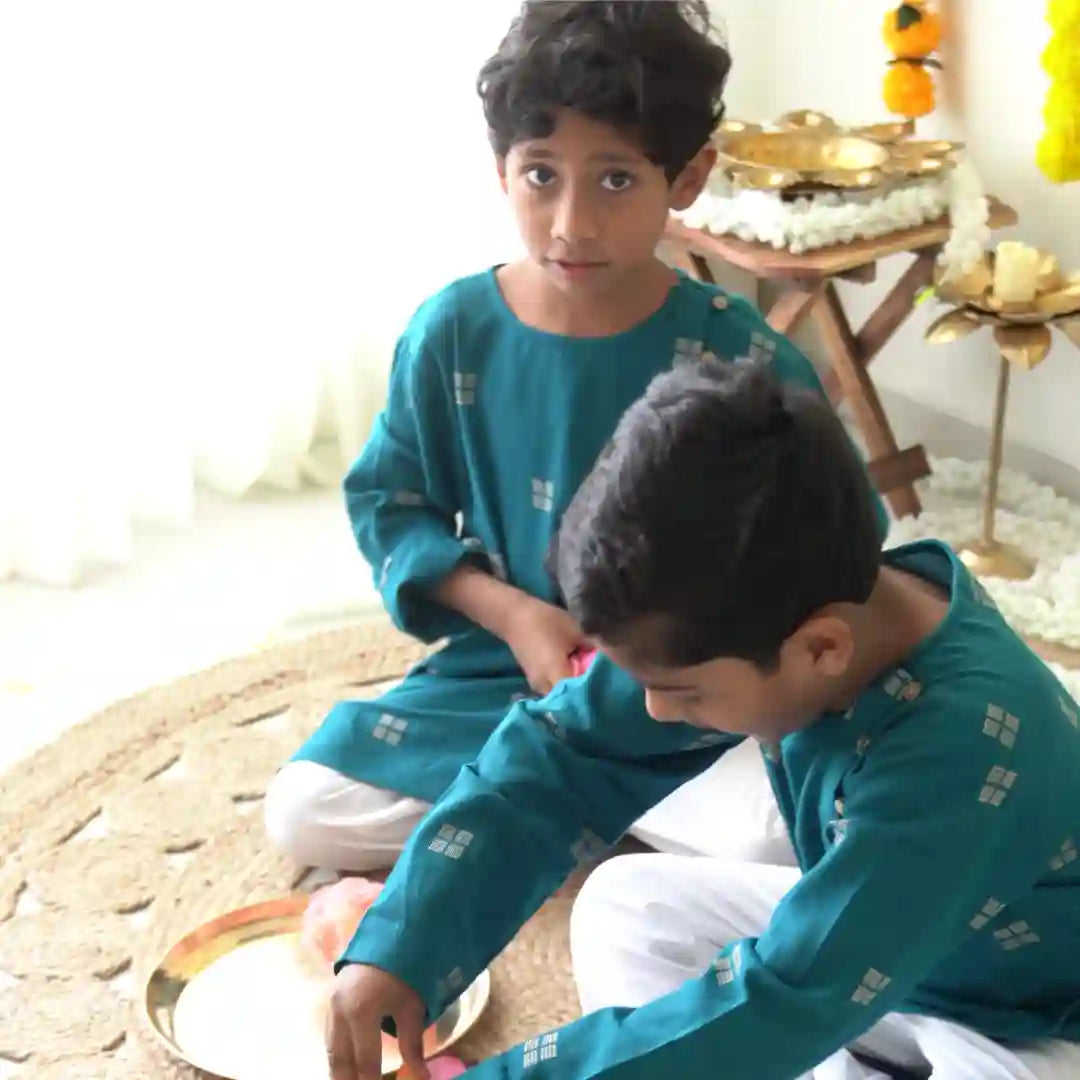 Two boys in teal kurtas and white pajamas sitting on a jute mat during festive preparations