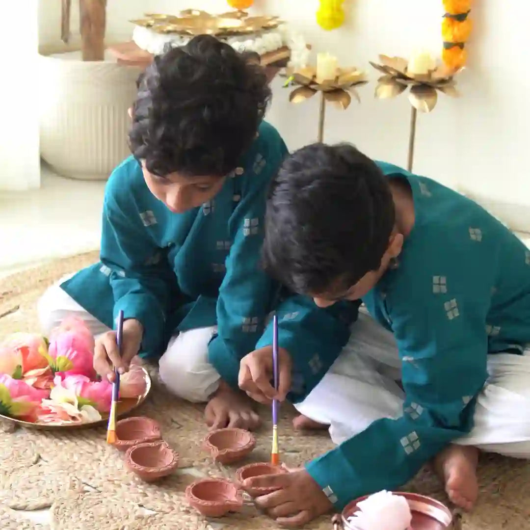 Two boys in teal handloom cotton kurtas painting clay diyas for a festive celebration